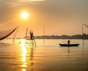 A fisherman rowing a small wooden boat past traditional lift nets on a river during a golden sunrise in Vietnam.