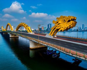 Aerial view of the famous Dragon Bridge in Da Nang, Vietnam, featuring a giant golden dragon head sculpture and yellow arches spanning the Han River under a clear blue sky with city skyline in the background
