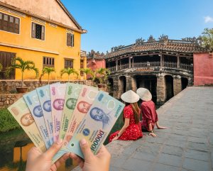 Hands holding Vietnamese Dong banknotes of 10,000, 20,000, 50,000, 100,000, 200,000 and 500,000 VND in front of a couple sitting by the canal near the Japanese Covered Bridge in Hoi An, Vietnam under a clear blue sky