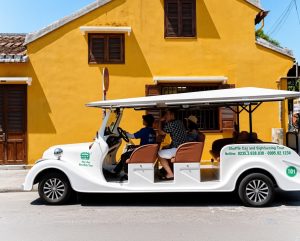 White open-air electric shuttle vehicle with brown seats carrying three passengers and a driver, parked in front of a bright yellow building with wooden shutters in Hoi An, Vietnam