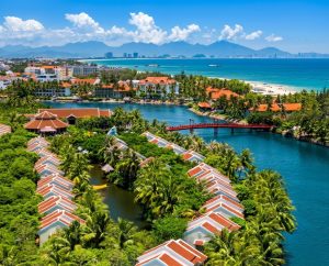 Aerial view of a luxury tropical resort in Hoi An surrounded by lush palm trees, a red bridge over a blue river, and coastal mountains under a sunny sky with white clouds